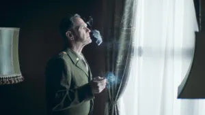 Een foto van een man in groen uniform, hij staat in een donkere kamer voor een raam een sigaret te roken.