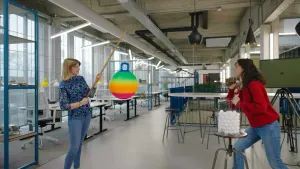 Een foto van een vrouw die een veelkleurige skippybal aan een hengel vasthoudt. Presentatrice Serah gooit pingpongballen naar de skippybal.