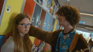 Meisje staat tegen kluisjes aan en kijkt ongemakkelijk in camera. Jongen heeft hand tegen kluisjes gezet en kijkt verliefd naar het meisje. 