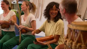 Een foto van presentatrice Serah. Ze zit tussen een rijtje muzikanten in een oefenzaaltje. Ze hebben allemaal traditionele instrumenten vast, eentje lijkt op een vishengel met een kokosnoot aan het handvat vast.