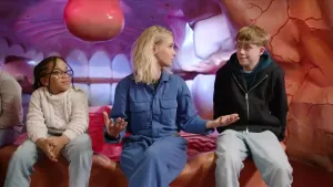 Janouk zit met twee kinderen die een metabole ziekte hebben in het lichaamsmuseum (corpus). Ze zitten voor een roze achtergrond die eruit ziet als de mond van binnen.