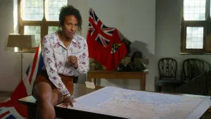 Presentatrice Astrid zit op de hoek van een tafel. Op tafel ligt een kaart en achter haar hangt een vlag.