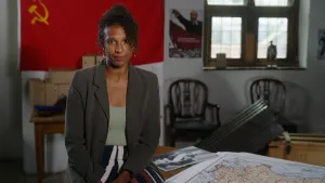 Presentatrice Astrid zit op een tafel op een oude zolder. Naast haar ligt een kaart en achter haar hangt een rode vlag.