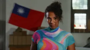 Presentatrice Astrid draagt een opvallend t-shirt met allerlei kleuren. Ze kijkt in de camera, staat op een zolder en achter haar hangt een vlag.