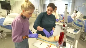 Janouk staat in het ziekenhuis met een studente naast zich. Ze hebben handschoentjes aan en oefenen op een kunstarm met bloedprikken.