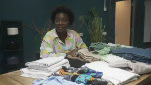 Een foto van presentatrice Tirsa die aan een tafel zit, daarop liggen allerlei kledingstukken en andere dingen van katoen.