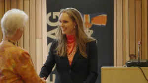 Een foto van Tjarda Struik, een blonde vrouw in een nette blazer met een grote burgemeestersketting om. Ze schudt een oudere vrouw de hand. Op de achtergrond staat een spreekgestoelte.