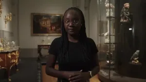 Thirsa staat in een ruimte met schilderijen en vitrines om zich heen. Er zijn veel gouden details achter haar te zien.