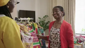 Presentator Thirsa staat samen met de Surinaamse oma Ria bij een kledingrek. Het rek is gevuld met kleurrijke traditionele kleding.