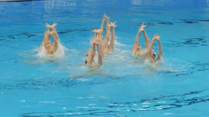 Een foto van een groep zwemmers in een zwembad. Ze staan in een rondje bij elkaar en steken allemaal hun beide armen sierlijk boven het water uit.