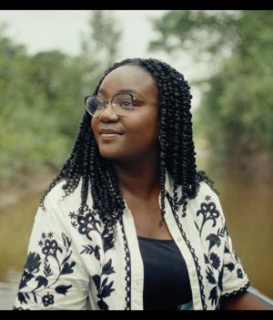 Een foto van presentator Tirsa in Suriname, ze is op het water, achter haar zie je een rivier en begroeiing op de kanten.