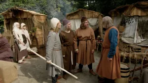 Foto van mensen in ouderwetse kleding voor hutjes op zandgrond.