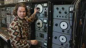 Een foto van presentator Pascal bij radio-apparatuur uit de jaren zestig. Op de apparatuur zitten knoppen en metertjes die waardes aangeven. Pascal draait aan een knop.