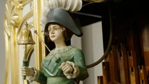 Foto van mannetje met hoed en bel op draaiorgel, houten buizen en goudkleurige versiering zijn zichtbaar op de achtergrond.