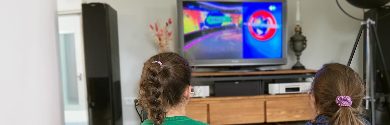 Foto van twee meisjes met bruin haar, gezien vanaf de rug, die op televisie naar het Jeugdjournaal kijken.