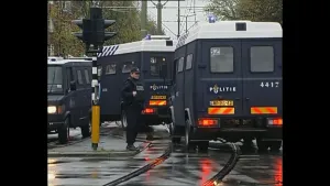 Een straat waarop drie donkerblauwe politiebusjes te zien zijn. In het midden staat een politieagent.