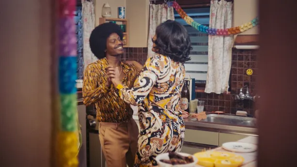 Een foto van een man en een vrouw die dansen in de keuken, ze hebben jaren 70 kleding aan en je ziet slingers hangen.