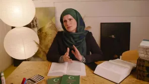 Er is een gesluierde vrouw te zien. Ze zit achter een tafel en heeft dikke boeken voor zich.