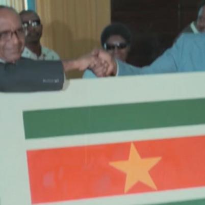 Een foto van twee mannen die elkaar de hand schudden voor zich houden ze een afbeelding van de vlag van Suriname.