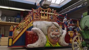 Presentator (vrouw) staat op een kleurrijke praalwagen. Er staat een prins op de wagen afgebeeld.