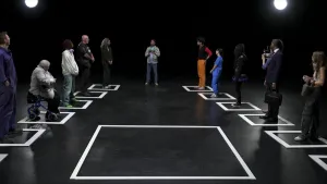 Een foto van een zaal. Op de grond zijn met tape hokjes afgeplakt. Allerlei verschillende mensen staan in de hokjes, zoals een oude vrouw met een rollator, een man in pak en een jonge alternatief geklede vrouw. In het midden is ook een groot leeg hokje.