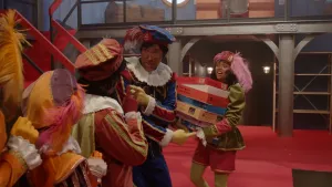 Een Piet staat met een stapel puzzeldozen. Hoofdpiet is in gesprek met andere Pieten.