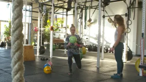 Een foto van presentator Janouk in de sportschool. Ze doet een sportoefening (lunge) terwijl ze een kettlebell vast heeft. Een trainer staat naast haar te kijken.
