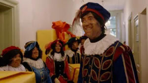 Hoofdpiet staat in de gang van Het Grote Pietenhuis. Om hem heen staan Pieten in een rij.