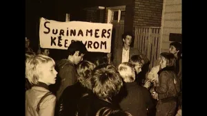 Foto van een groep mensen die buiten staan. Iemand houdt een spandoek omhoog met daarop 'Surinamers keer om'.