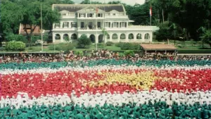 Still uit archiefbeeld waarop inwoners van Suriname op een groot plein de vlag van Suriname vormen: groen, wit en rood met een gele ster in het midden.