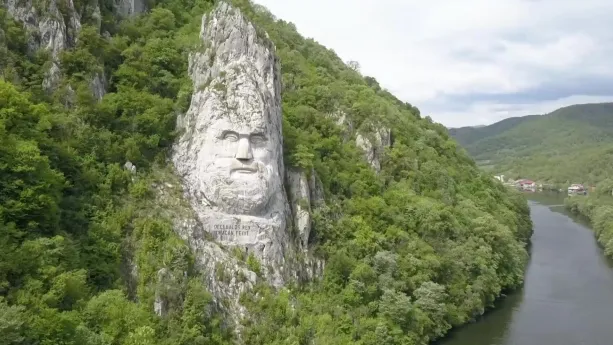 Een foto van een Roemeens landschap met links van de rivier een uit de rotswand uitgehouwen hoofd van een koning van de Daciers.