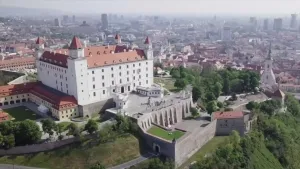 Een foto van de Slowaakse hoofdstad Bratislava met op de voorgrond een groot wit kasteel, daarachter de rest van de stad met hoge gebouwen.