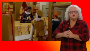 Hoofdpiet spreekt Pieten toe in de Pakjeskamer. Ze zijn omringd door noodpakketten. Gebarentolk staat naast het scherm.