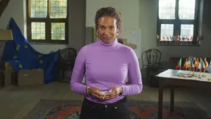 Presentatrice Astrid staat op een zolder. Naast haar een EU vlag en een tafel met vlaggetjes erop.