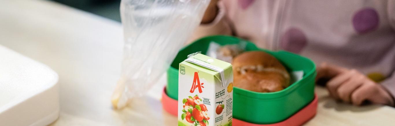 Foto van broodtrommel met een broodje op tafel en een pakje appelsap en leeg boterhamzakje ernaast.
