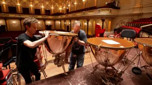Foto van twee mannen die een zware trommel een podium optillen in een concertzaal.