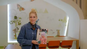 Een foto van presentatrice Janouk met in haar handen een foto, ze staat voor een tafel met stoelen.