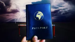 Foto van hand die blauw paspoort vasthoudt, op de achtergrond een roodwitte slagboom, witte wolken en een windmolen.