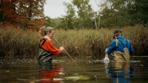 Foto van presentatrice Petra Grijzen samen met een wetenschapper in een sloot om te zoeken naar methaan.