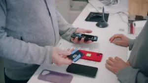 Een foto van twee mensen aan weerszijden van een balie, je zet alleen hun buik en handen, op de balie liggen een aantal mobieltjes. De persoon links houdt een telefoon vast die opengemaakt is en in zijn andere hand de batterij.