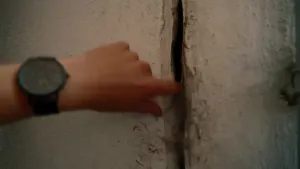 Een foto van een hand met een groot horloge om de pols die een brede scheur in een muur aanwijst, de hand gaat met 1 vinger in de scheur.