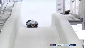 Foto van skeletonatleet die liggend op zijn buik op een slee van een bevroren bobsleebaan suist. Hij draagt een witte helm en een lichtblauw pak.