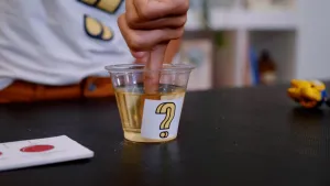 Foto van een vinger in een bekertje gele vloeistof met een vraagteken erop.