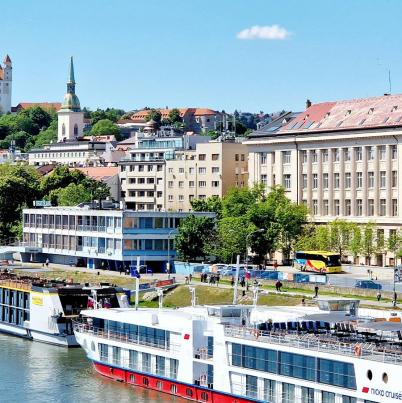 Foto van Bratislava met gebouwen aan het water. Aan de kade liggen boten. In de verte staat een kasteel op een berg.