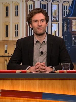 Foto van presentator Roel Maalderink achter de desk van Kiespijn, grote rode potloden liggen voor hem en achter hem een backdrop van het torentje in Den Haag.