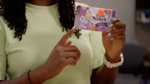 Foto van presentatrice die verpakkingsdoosje van broodbeleg mini's vasthoudt, een kartonnen doosje met paarse opdruk.