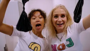 Foto van presentatrices Janouk en Serah met zwarte plastic zakjes met daarin verse hondendrollen. Ze kijken jolig in de camera en staan voor een wc.