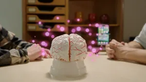 Foto van tafel met een wit model van hersenen waar roze stipjes omheen zweven. Op tafel liggen de armen van twee mannen die met elkaar in gesprek zijn over smartphonegebruik.