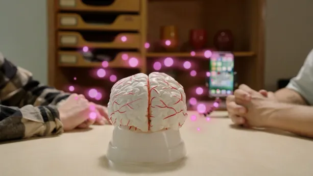 Foto van tafel met een wit model van hersenen waar roze stipjes omheen zweven. Op tafel liggen de armen van twee mannen die met elkaar in gesprek zijn over smartphonegebruik.