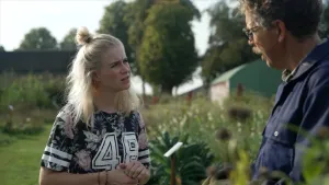 Foto van Klokhuispresentatrice Janouk met een boer in een blauwe overall tussen zijn vergeten groenten.